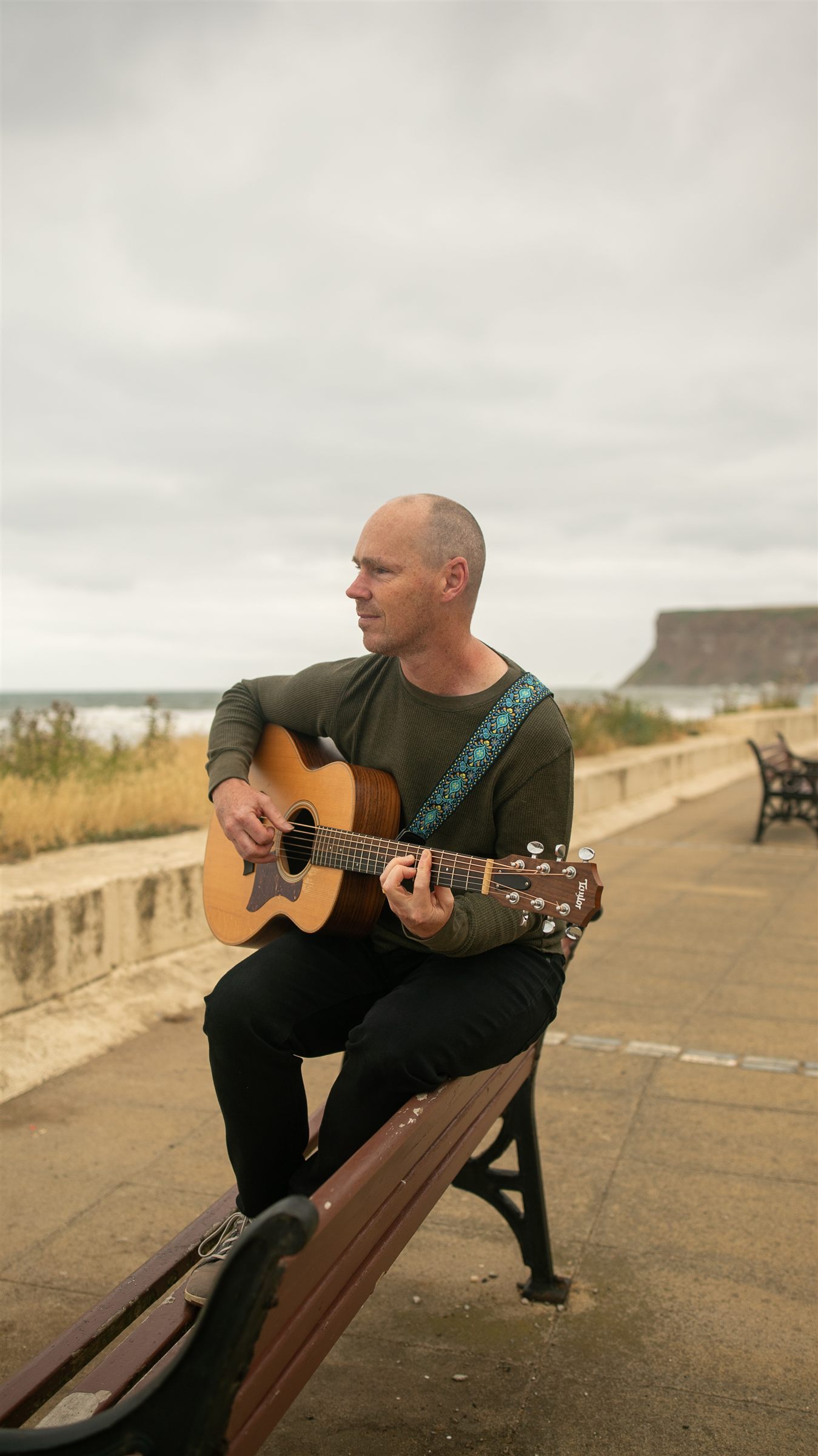 John Grummitt sitting on a seafront bench with his guitar